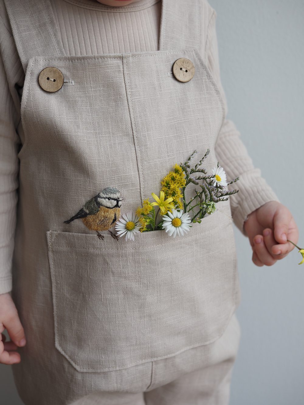 Helle Latzhose mit gestickter Blaumeise und Frühlingsblumen in der Tasche