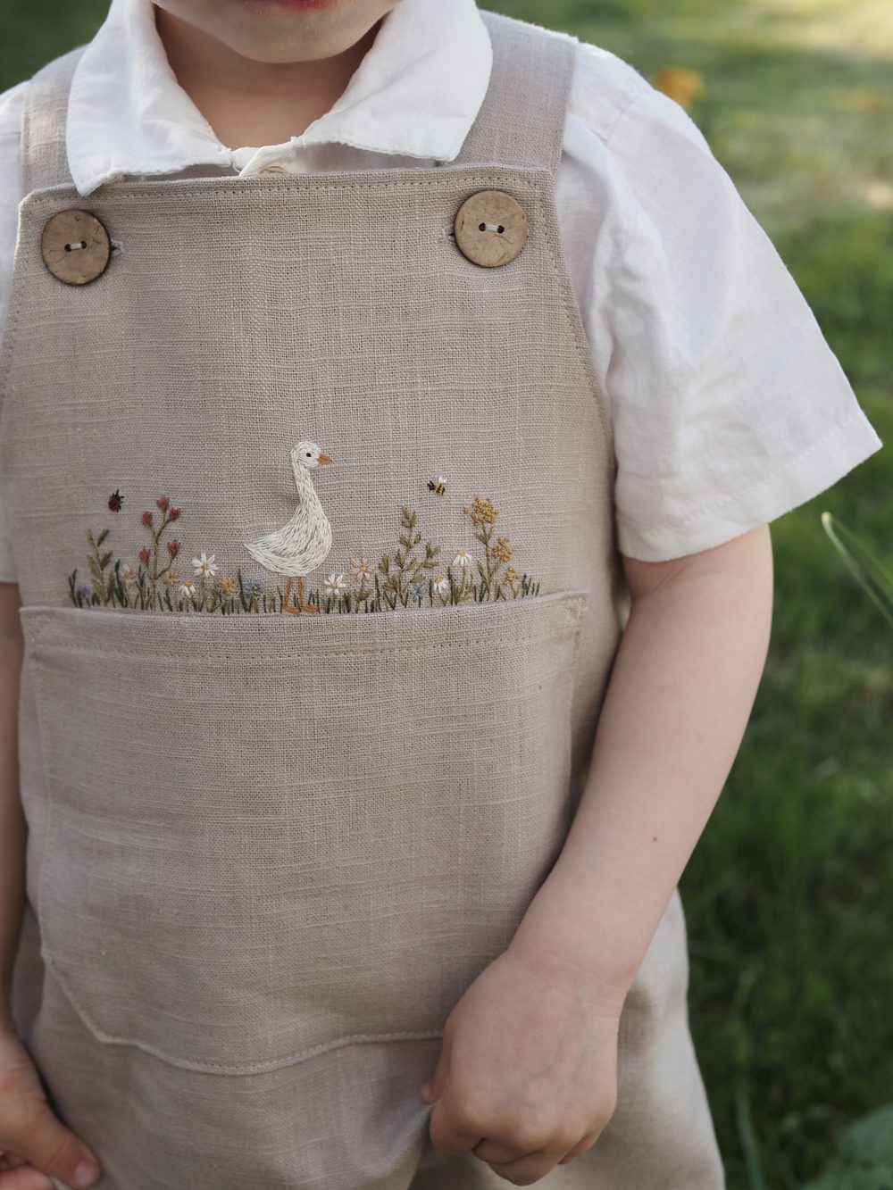 Kind in heller Leinenlatzhose mit gestickter Gans, Löwenzahn in der Hand