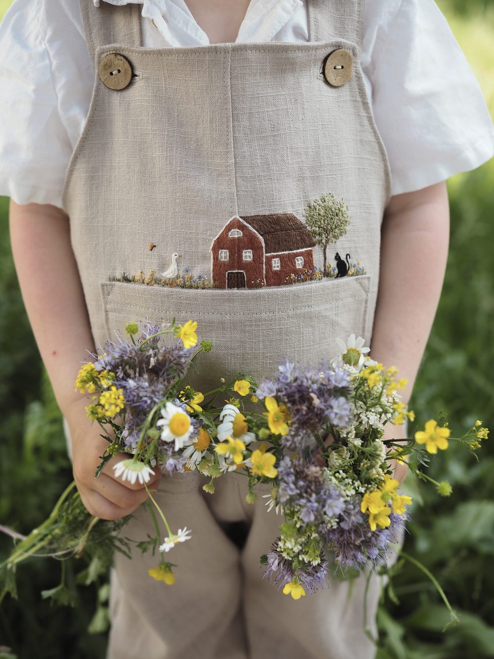 Kind mit Blumenstrauß in bestickter Latzhose mit Häuschenmotiv